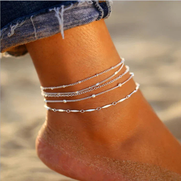 Close-up of ankle wearing layered delicate anklets, neutral background. Gold and silver chains, beaded details. Soft lighting, minimalist styling, professional jewelry photography